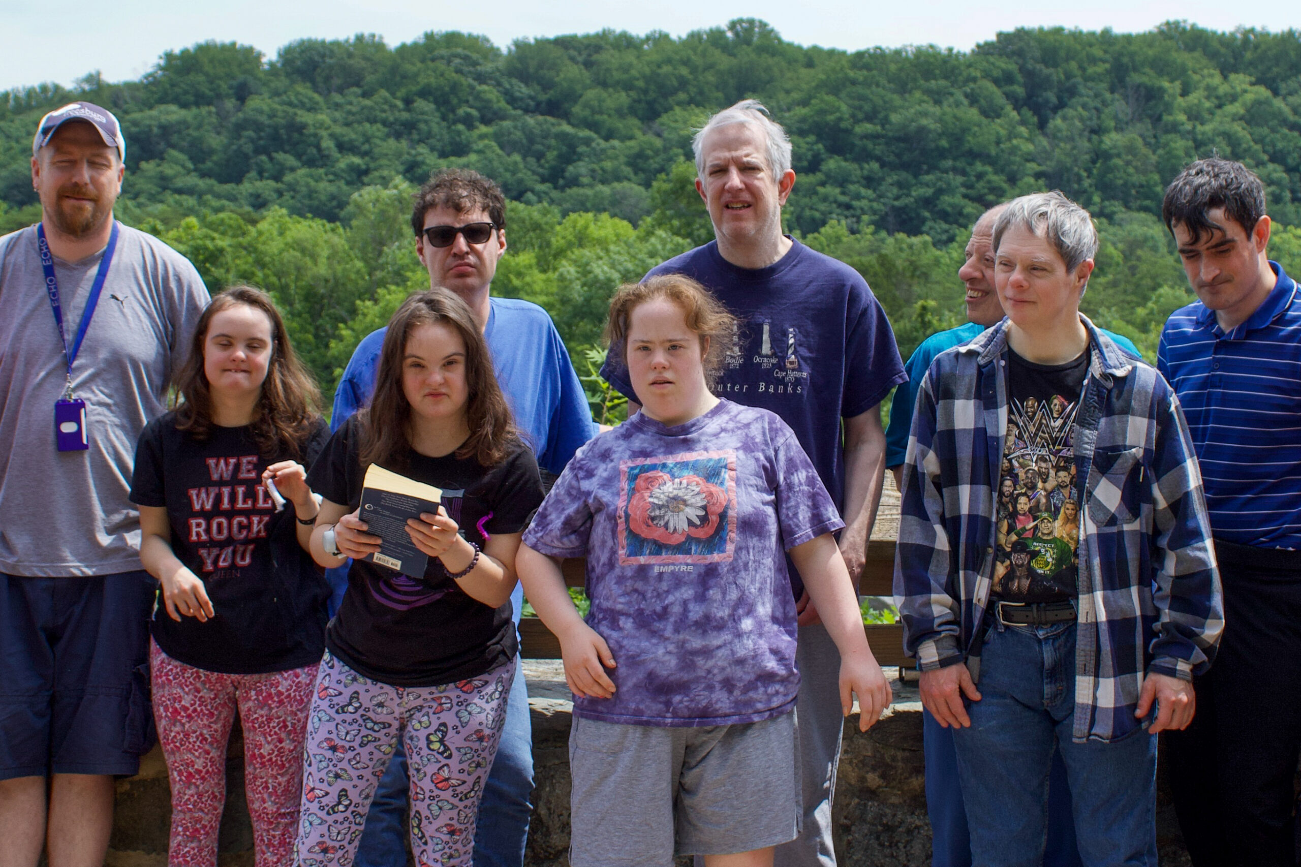 A group of nine people, including men and women of various ages, stand outdoors in front of a stone railing with a forested hillside in the background. Some are smiling, and one person holds a book. The group is casually dressed in T-shirts and shorts or jeans.<br />
